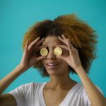 Smiling woman with afro hair holding cryptocurrency coins in front of eyes, against blue background.