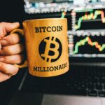 Golden mug with 'Bitcoin Millionaire' text in front of stock market charts on laptop screen.