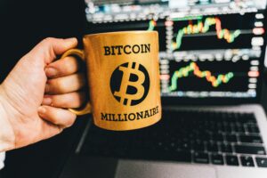 Golden mug with 'Bitcoin Millionaire' text in front of stock market charts on laptop screen.